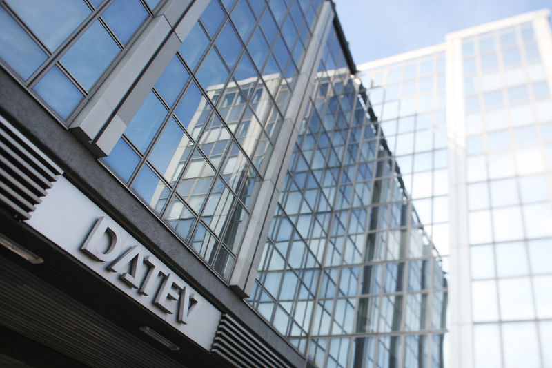 DATEV-Bürogebäude mit dominanter Glasfassade und moderner Stahlrahmenstruktur. Die Perspektive zeigt den Eingangsbereich von schräg unten, wodurch die Höhe des Gebäudes betont wird. Spiegelnde Fenster und ein klarer Himmel sorgen für einen transparenten, offenen Eindruck