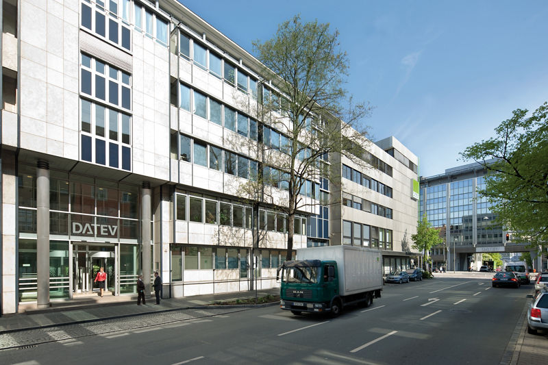 DATEV-Verwaltungsgebäude mit hell verkleideter Fassade und dem grün-weißen Logo über dem Eingangsbereich. Der Blick fällt entlang einer städtischen Straße mit parkenden Autos. Fußgänger bewegen sich vor dem Hauptportal. Die Architektur ist funktional und geschäftlich geprägt.