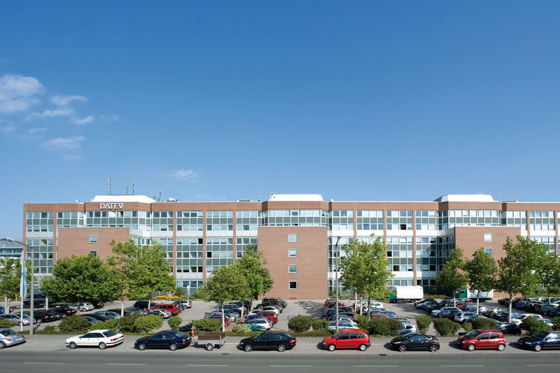 Großes DATEV-Gebäude mit rotbrauner Klinkerfassade, flachem Dach und weißen Fensterrahmen. Davor liegt ein weitläufiger Parkplatz mit zahlreichen Autos und Bäumen. Das Gebäude wirkt funktional und ist in ein urbanes Umfeld eingebettet. Ein klarer, blauer Himmel rundet das Bild ab