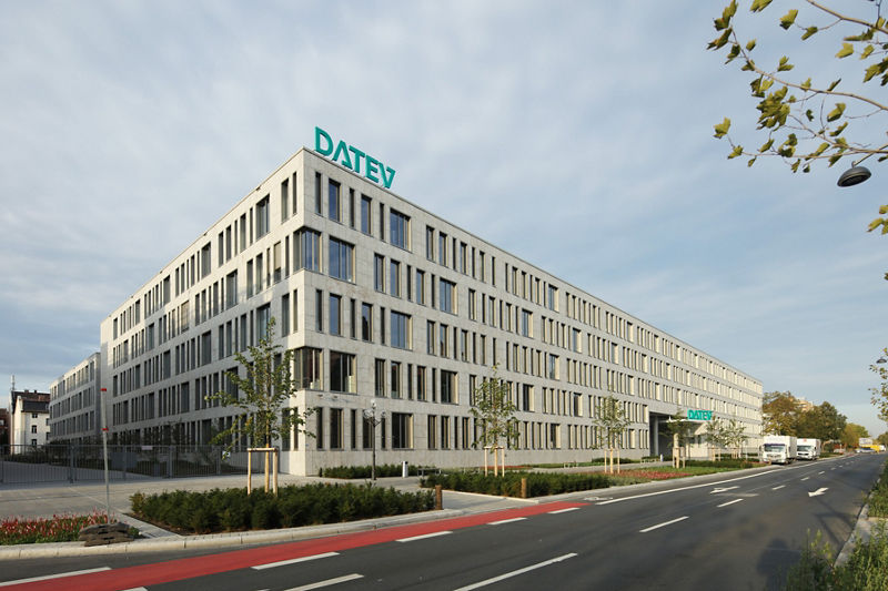 Panoramaansicht des modernen DATEV IT-Campus in Nürnberg. Das mehrstöckige Bürogebäude mit heller Steinfassade erstreckt sich entlang einer breiten Straße. Auf dem Dach prangt gut sichtbar das grüne DATEV-Logo. Junge Bäume säumen den Gehweg, auf der Straße parken Transporter. Die Szenerie vermittelt Weitläufigkeit und Offenheit des Firmensitzes.