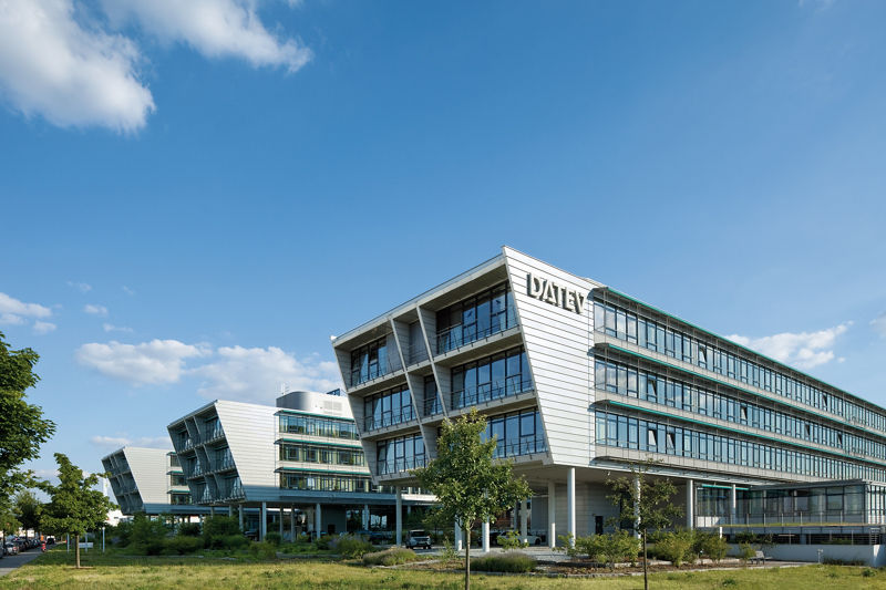 Weitläufige Aufnahme eines architektonisch markanten DATEV-Gebäudes mit mehrfach gestaffelten Glastrakten. Die moderne Fassade ist geprägt von horizontalen Linien, die dynamisch wirken. Der grüne DATEV-Schriftzug ist gut sichtbar am Gebäude angebracht. Im Vordergrund erstreckt sich eine begrünte Fläche mit Bäumen unter blauem Himmel.