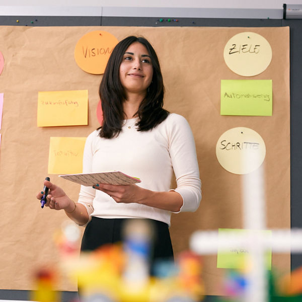 Frau steht vor einer Pinnwand mit bunten Notizzetteln und Stichworten wie ‚Vision‘, ‚Ziele‘ und ‚Schritte‘, hält einen Block und einen Stift in der Hand.