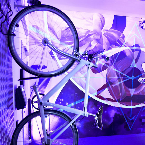 Weißes Fahrrad hängt senkrecht an einer Wandhalterung; im Hintergrund ein buntes Wandgemälde in Lila- und Blautönen mit geometrischen Mustern.