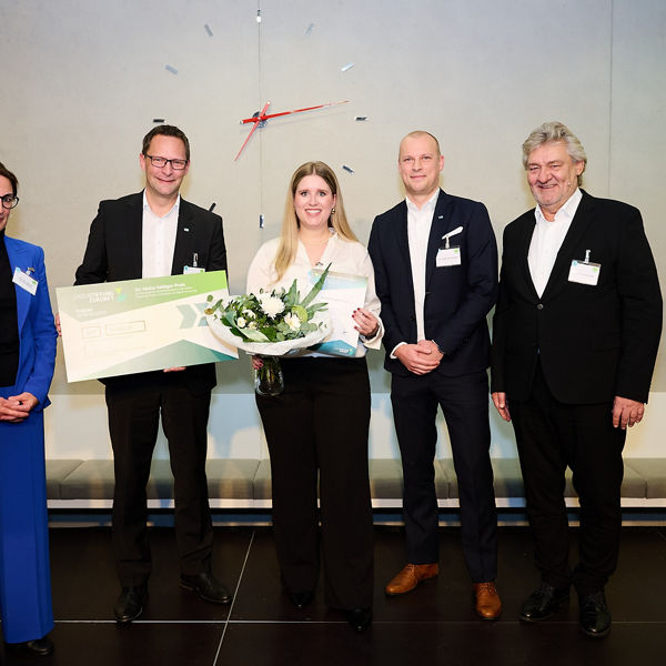 Fünf Personen stehen nebeneinander bei der Preisverleihung des Dr.-Heinz-Sebiger-Preises 2024. Eine Frau in der Mitte hält einen Blumenstrauß und eine Urkunde, daneben überreicht ein Mann einen symbolischen Scheck über 10.000 Euro von der DATEV-Stiftung Zukunft. Alle sind festlich gekleidet und lächeln in die Kamera.