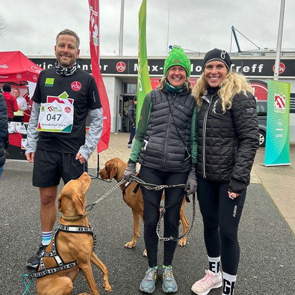 Drei Teilnehmende des Spendenlaufs vor dem Max-Morlock-Stadion lächeln in die Kamera; zwei Hunde an der Leine sitzen und stehen neben ihnen.