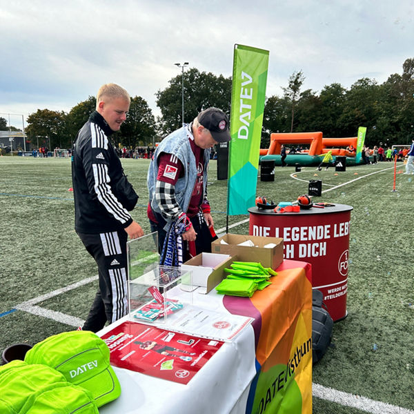 Zwei Personen an einem DATEV-Stand mit grünen Caps, Flyern und Werbematerial auf einem Fußballfeld bei der Bayerischen Inklusionsfußballmeisterschaft. Im Hintergrund aufblasbare Spielgeräte.