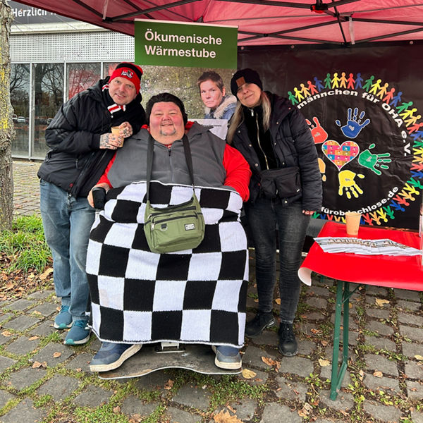 Vier Personen unter einem roten Pavillonzelt, eine davon im Rollstuhl mit Decke; Schild ‚Ökumenische Wärmestube‘ hängt über ihnen.