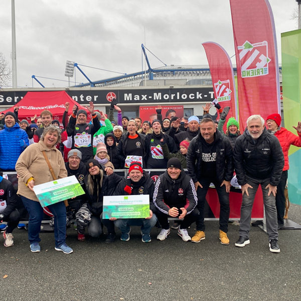 Gruppe von Menschen in Sportkleidung und Winterjacken posiert jubelnd vor dem Max-Morlock-Stadion; im Vordergrund werden große Spendenschecks hochgehalten, daneben bunte DATEV- und Vereinsfahnen.