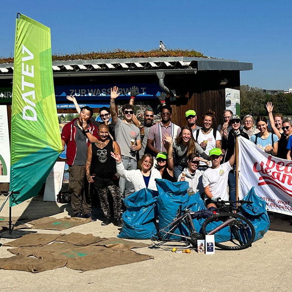 Gruppe von Menschen posiert fröhlich im Freien mit DATEV-Fahnen und einem Banner ‚Beach Cleaner‘; im Vordergrund liegen gesammelte Müllsäcke und Fahrräder.
