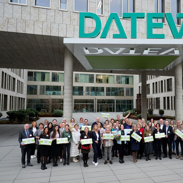 Gruppenfoto vor dem DATEV-Hauptgebäude in Nürnberg mit über 50 Personen, die symbolische Spendenschecks halten. Die Spendenempfänger:innen stehen gemeinsam unter dem großen DATEV-Schriftzug im Eingangsbereich und lächeln in die Kamera.