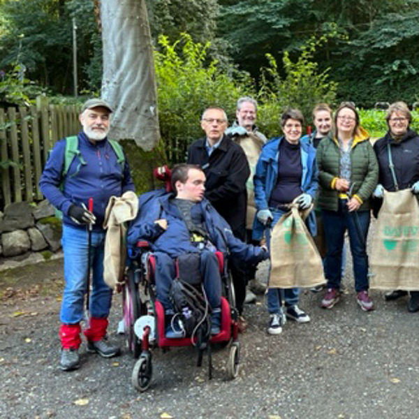 Gruppe von Menschen mit Handschuhen und Jutesäcken posiert bei einer Aufräumaktion im Wald; vorne ein Mann im Rollstuhl und ein kleiner weißer Hund.