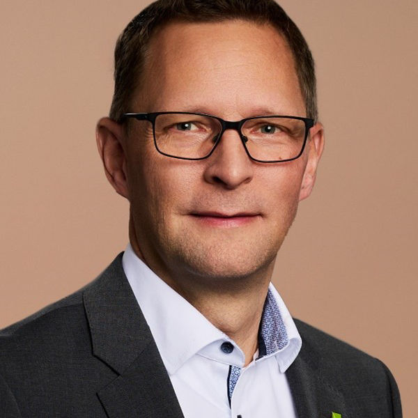 Portrait von Dr. Markus Algner, er trägt einen dunklen Anzug und ein weißes Hemd mit offenem Kragen vor beigem Hintergrund.