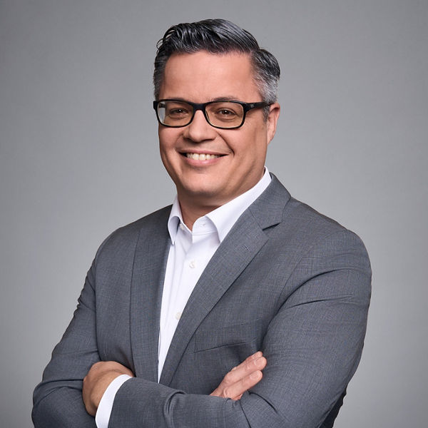 Portrait von Markus Munz in grauem Sakko und weißem Hemd vor grauem Hintergrund.