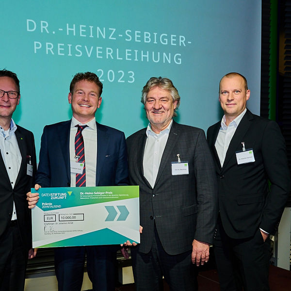 Vier Männer in Anzügen stehen vor einer Leinwand mit dem Schriftzug ‚Dr.-Heinz-Sebiger-Preisverleihung 2023‘. Einer von ihnen hält einen symbolischen Scheck in Höhe von 10.000 Euro von der DATEV Stiftung Zukunft. Alle blicken lächelnd in die Kamera.