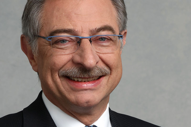 Porträt eines älteren Mannes mit grauen Haaren, Brille und Schnurrbart. Er trägt einen dunklen Anzug mit weißem Hemd und Krawatte und einer DATEV-Anstecknadel. Er lächelt freundlich in die Kamera. Der Hintergrund ist neutral und hellgrau.