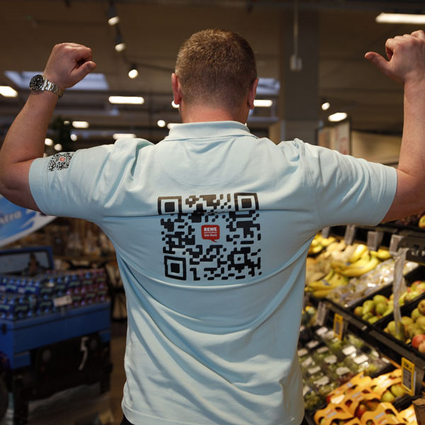 Ein Mann steht mit dem Rücken zur Kamera in einer Obstabteilung eines Supermarkts und flext die Arme. Auf seinem hellblauen Polo-Shirt ist ein großer QR-Code mit REWE-Logo aufgedruckt. Im Hintergrund Obstregale und Verkaufsfläche.