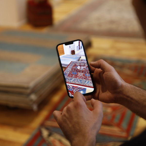 Eine Person hält ein Smartphone über einen hochwertigen Orientteppich und nutzt eine AR-Anwendung namens ‚sarfi‘ zur digitalen Erkennung oder Erfassung. Im Hintergrund liegen weitere Teppiche in einem Showroom.