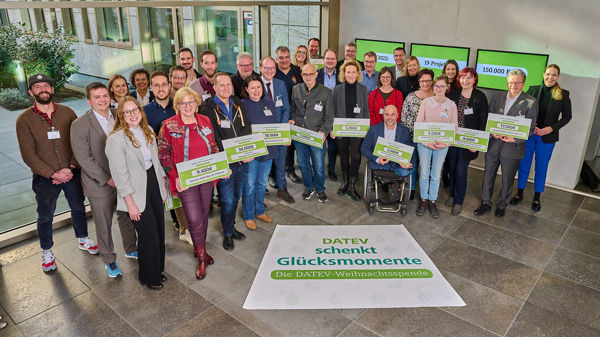 Gruppenfoto mehrerer Personen mit symbolischen Spendenschecks bei der Aktion ‚DATEV schenkt Glücksmomente – Die DATEV-Weihnachtsspende‘. Im Hintergrund sind Bildschirme mit Informationen zur Spendenaktion sichtbar. Die Gruppe steht im DATEV-Gebäude.